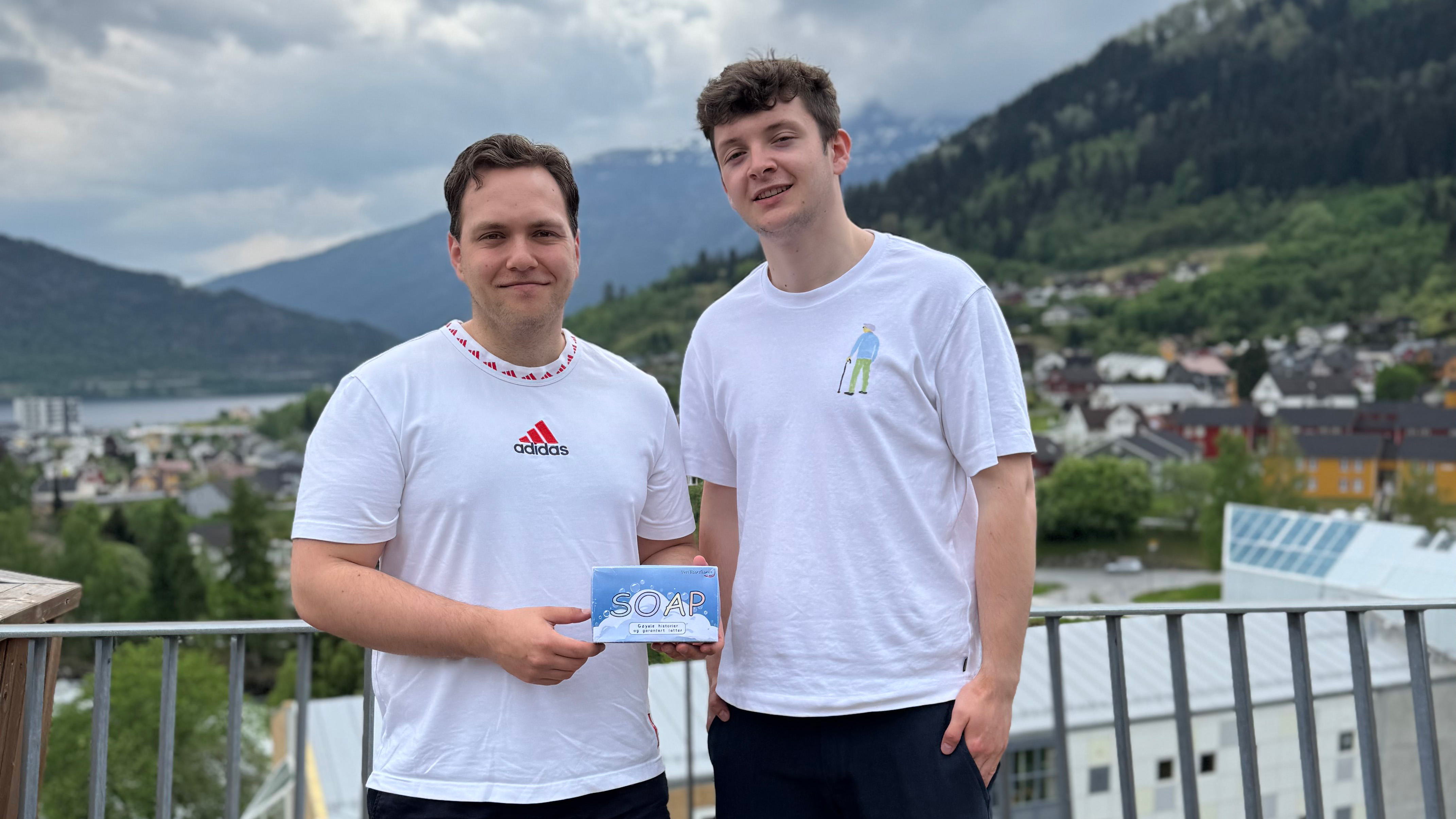Simen og Tommy, to karar med brunt hår, kledd i kvite T-skjorter og mørke bukser, viser smilande fram ein boks som inneheld brettspelet SOAP. I bakgrunnen ser ein fjell, fjord grå skyer og ymse hus.