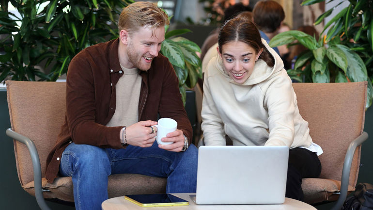 To studentar som smiler og sit i sofaen. Dei jobbar saman på ein PC.